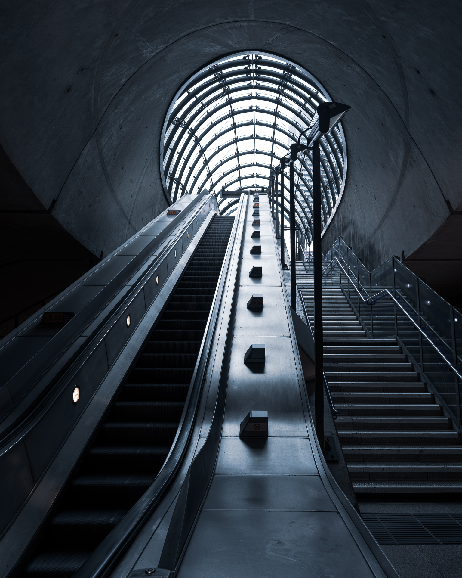 Escalators rise through a circular opening beneath the curved concrete architecture of Canary Wharf Underground station in London.